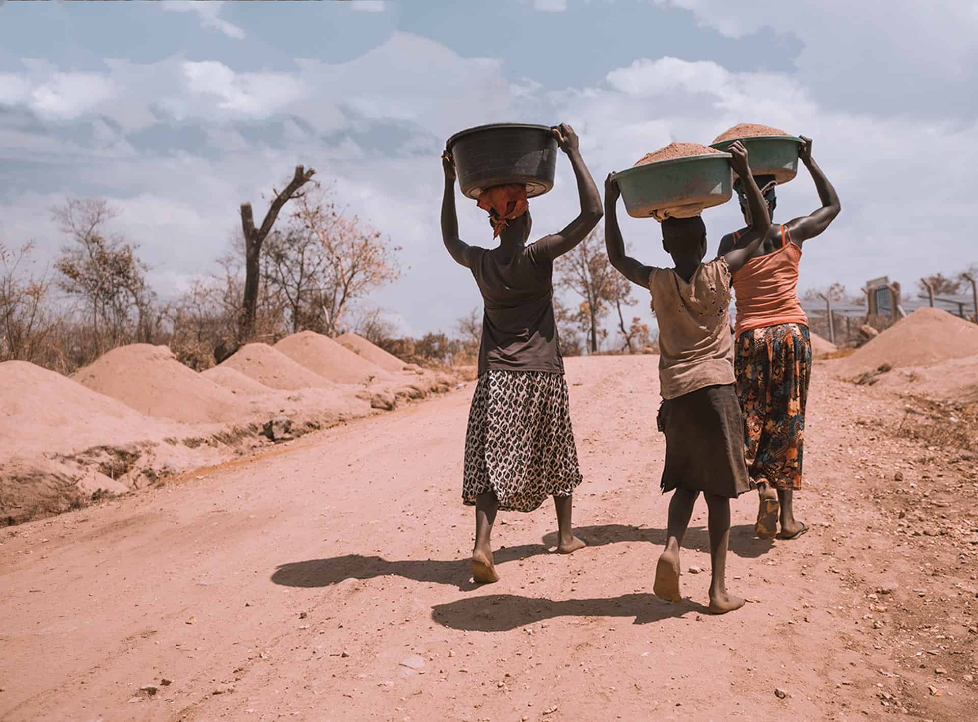 Three individuals carrying basins on their heads walking along a dirt road in a beautiful town.